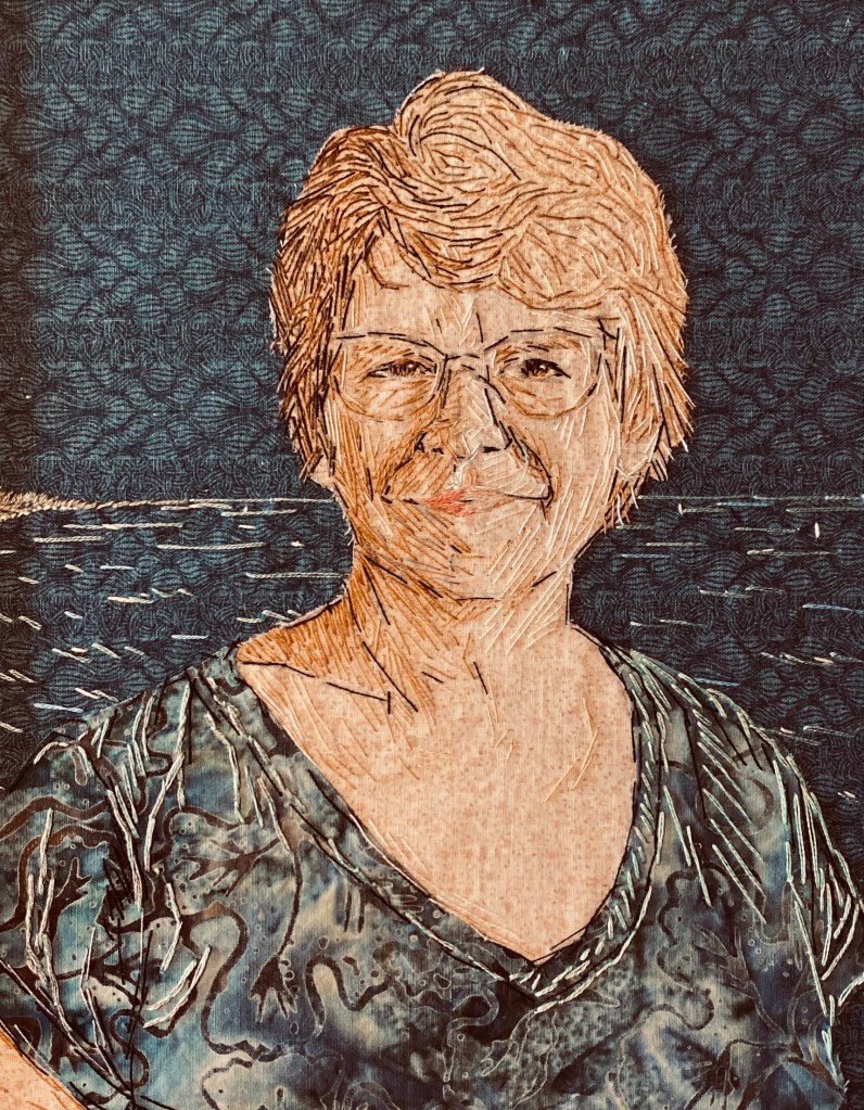 This artwork by Angela called 'Selfie Stitch' is a self portrait created with needlework. She is a white woman with cropped fair hair and glasses. 