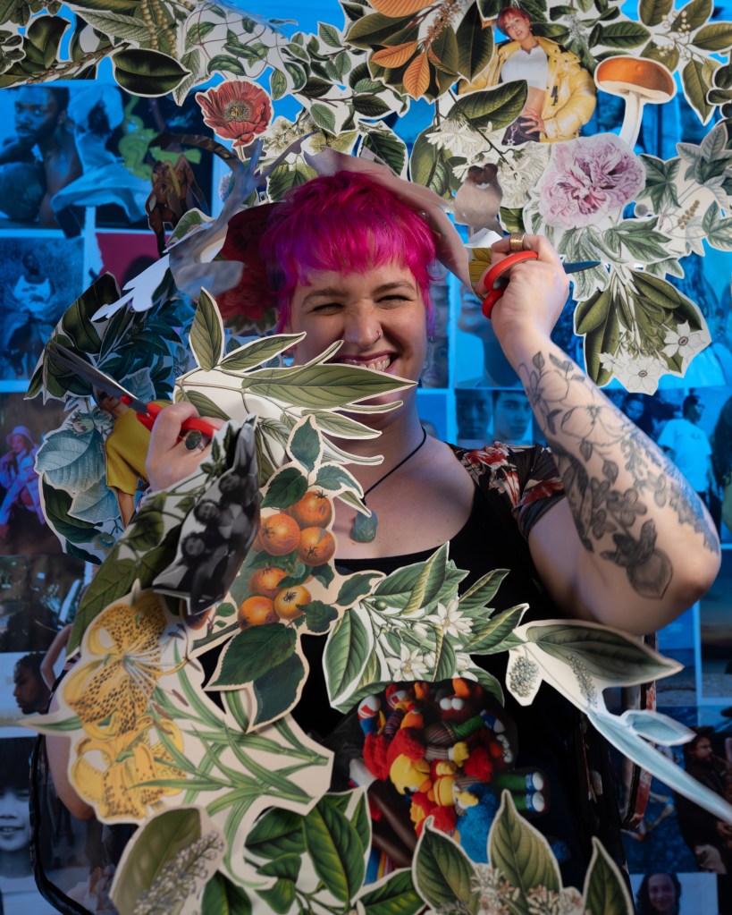 An artist photo: Emily is a white woman with pink hair and tattoos of fruit and flowers on her arm. She is bursting out of some paper collage materials. 