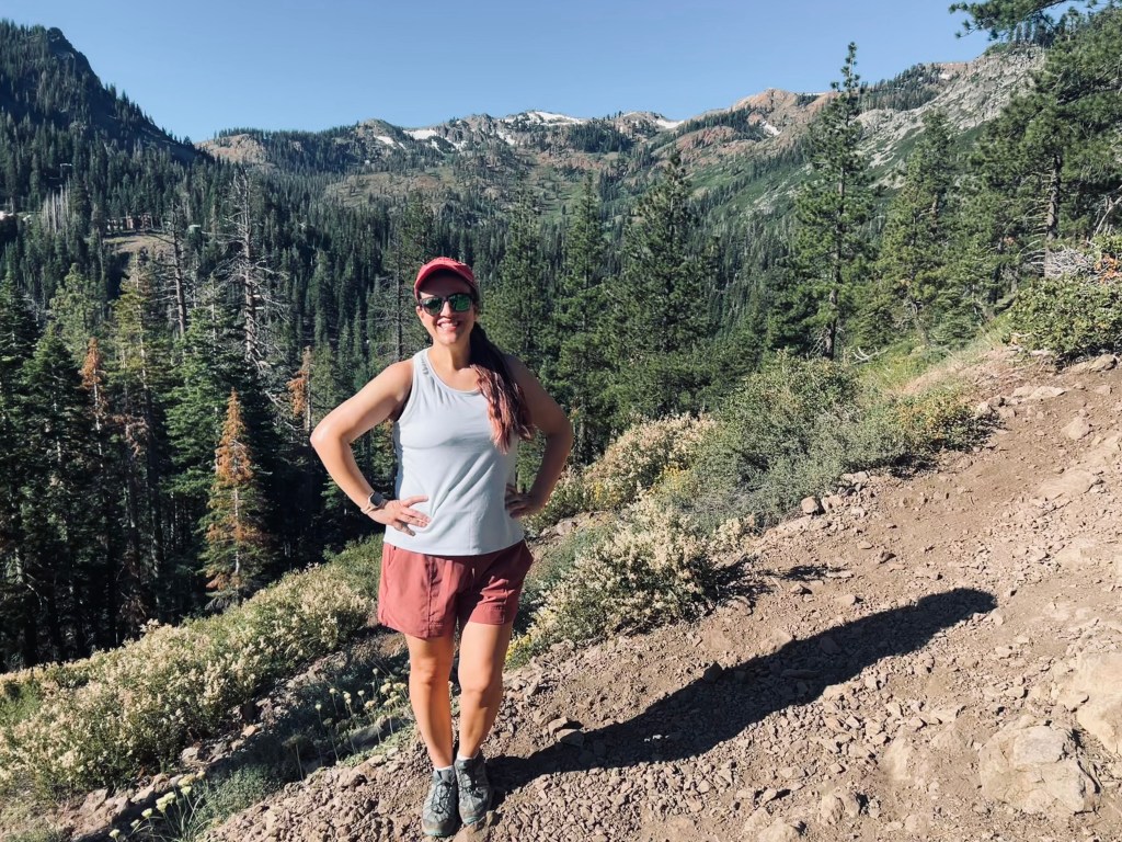Angie stands on a rugged path. She is a white woman with long brown hair wearing hiking clothes. Behind her forest and the tops of mountains. 