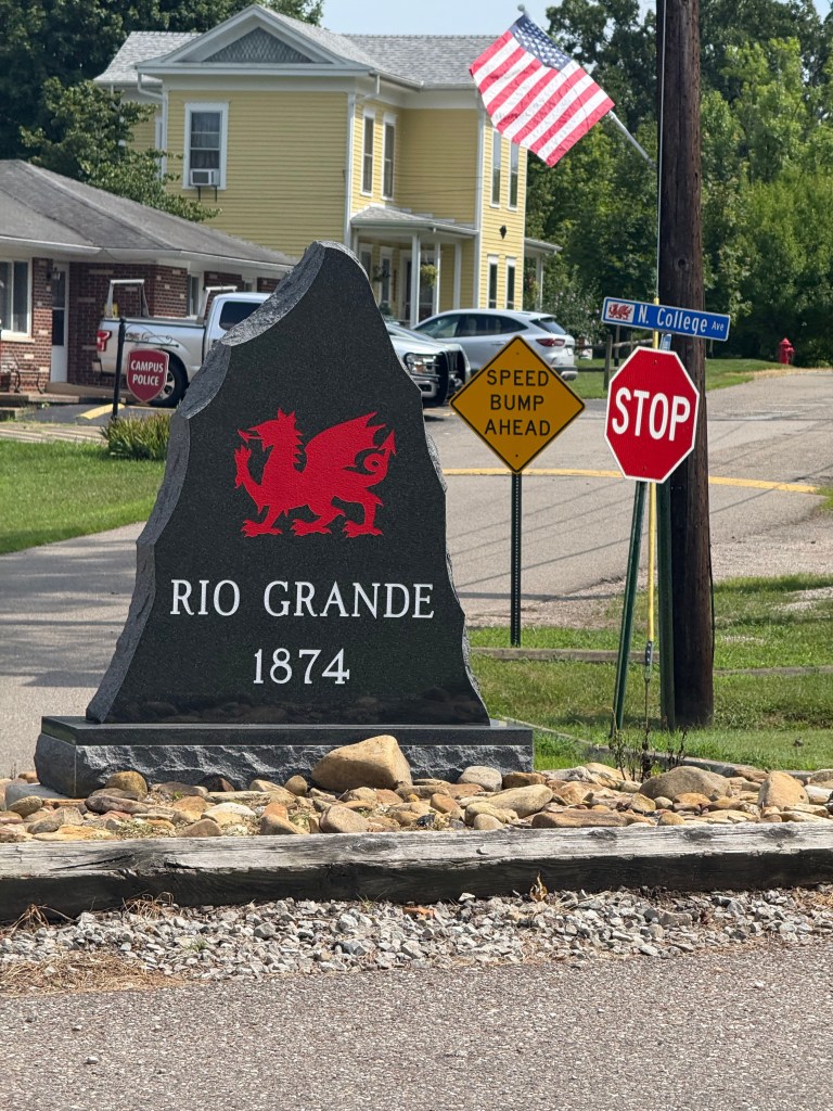 A welcome sign for Rio Grande, featuring a red dragon emblem and the year 1874, surrounded by rocks, with traffic signs including 'Speed Bump Ahead' and 'Stop' in the background.